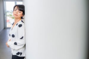 Adult woman in a stylish polka dot blouse with eyes closed, leaning against a white wall indoors.