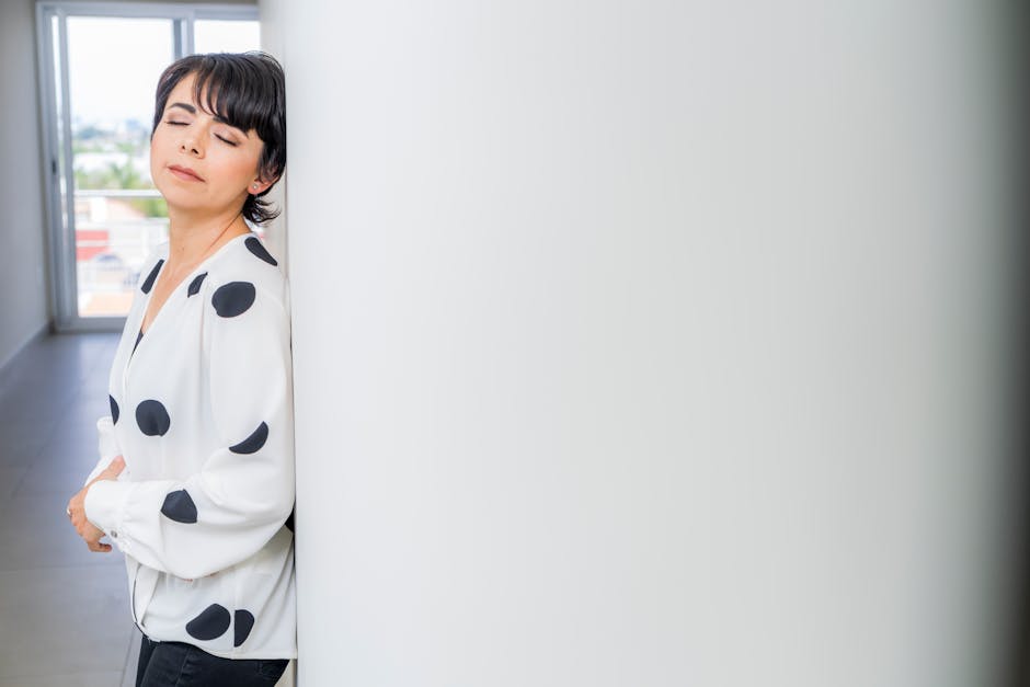 Adult woman in a stylish polka dot blouse with eyes closed, leaning against a white wall indoors.