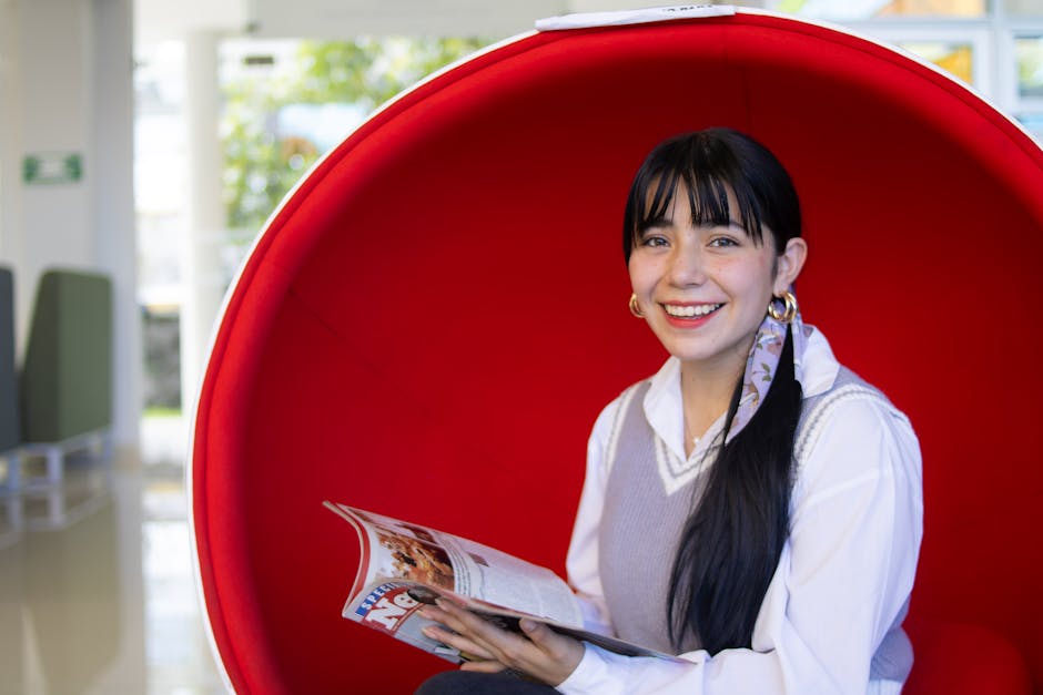 Smiling woman reading a magazine in a red chair, capturing a relaxed and modern indoor scene.