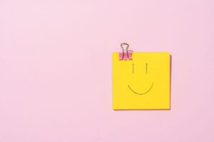 Yellow sticky note with a smiley face clipped to a pink surface, conveying happiness and simplicity.