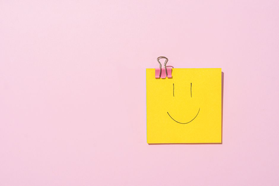 Yellow sticky note with a smiley face clipped to a pink surface, conveying happiness and simplicity.